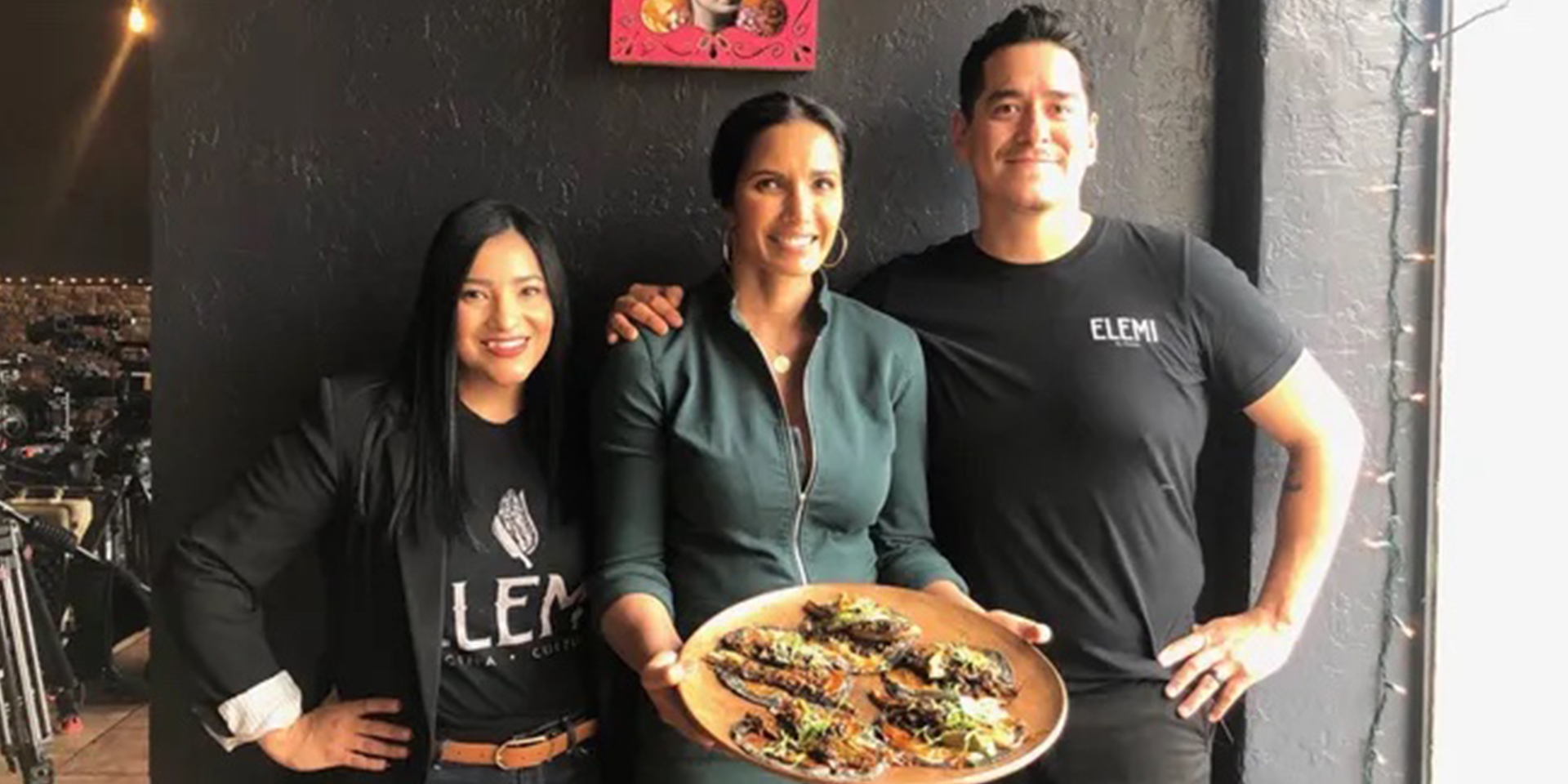 Smiling people standing in front of a black wall holding a plate with tacos.